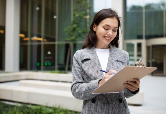 Smiling young businesswoman reading documents and signing papers on clipboard, conducting professional work standing outside in urban city area, writing business report