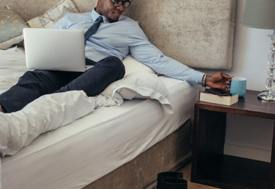 Confident man in formal clothes working on laptop while lying in bed. Man reaching out to coffee mug placed on table while relaxing on bed.