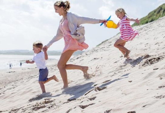 Young single parent family holding hands and running down the sanddunes to the sea. They are laughing and wearing casual clothing.